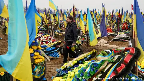 A man carries a flower as he walks through a cemetary full of Ukrainian flags