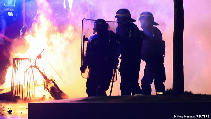 Drei Polizisten mit Schutzausrüstung stehen auf einer Straße und beobachten ein Feuer, in dessen Flammen ein Mülleimer und ein E-Roller zu sehen sind.