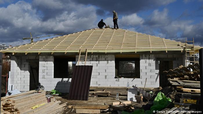 Ukraine Krieg | Wiederaufbau in Butscha, ein Haus im Rohbau mit Dachstuhl ohne Schindeln, zwei Männer auf dem Dach, einer hockt und einer steht, davor Baumaterial