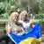 Irina Shevchenko (e.) e Alla Hrechana (d.) sentadas em banco em praça de São José dos Campos e segurando uma bandeira da Ucrânia.