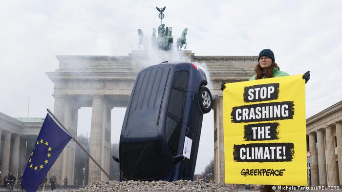 Автомобиль застрял капотом в куче гравия. Рядом с ним молодая женщина держит плакат Гринпис с надписью «Хватит разрушать климат» на фоне Бранденбургских ворот.
