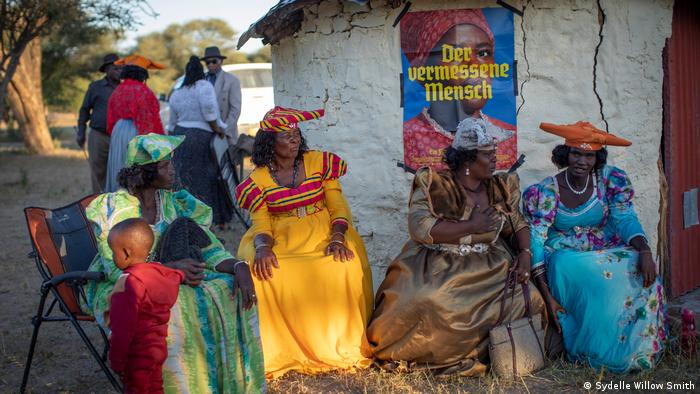 Frauen sitzen in farbigen Kleidern in einem Dorf vor einem Filmplakat zu Der vermessene Mensch.