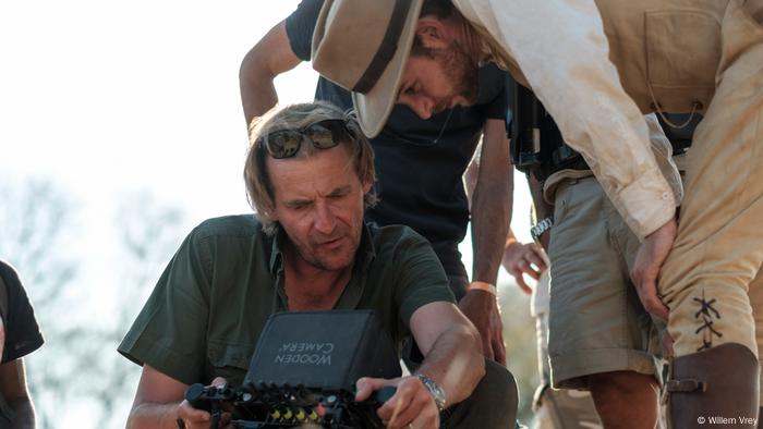 Regisseur Lars Kraume bei den Dreharbeiten in Namibia mit Sonnenbrille auf dem Kopf schaut auf den Screen einer Kamera. Hinter ihm beugt sich der Hauptdasteller in Kolonialoutfit über ihn