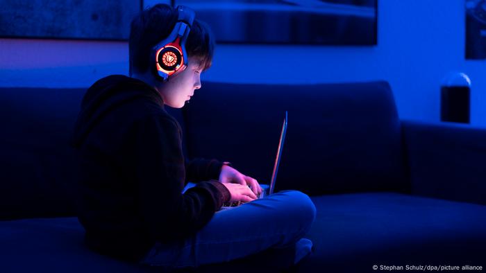 Ein Junge sitzt mit Kopfhörern und einem Laptop auf den Beinen in einem Zimmer in bläulichem Licht. 