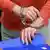 A imagem mostra as mãos algemadas do réu condenado por tentativa de homicídio de um policial na Alemanha. As mãos seguram uma pasta. O sujeito veste blusão vermelho.