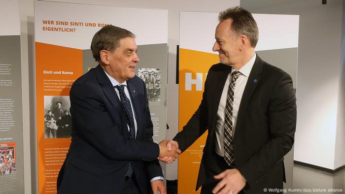 Zwei Männer im Hemd, Krawatte und dunklem Anzug geben sich die Hand. Im Hintergrund ist eine Stellwand mit Fotos zu erkennen