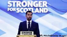 Ein Mann steht an einem Rednerpult vor einer Wand mit der Aufschrift Stronger for Scotland. Er trägt einen Anzug und spitzt seine Lippen.