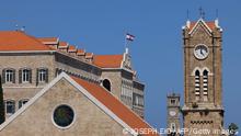 Die Spitze eines Kirchturms zeigt eine Uhr mit der Zeit 12:25 Uhr. Auf dem Dach des Gebäudes daneben weht eine Flagge mit einem grünen Stern auf weißem Hintergrund in der Mitte, oben und unten ist ein roter Balken. 