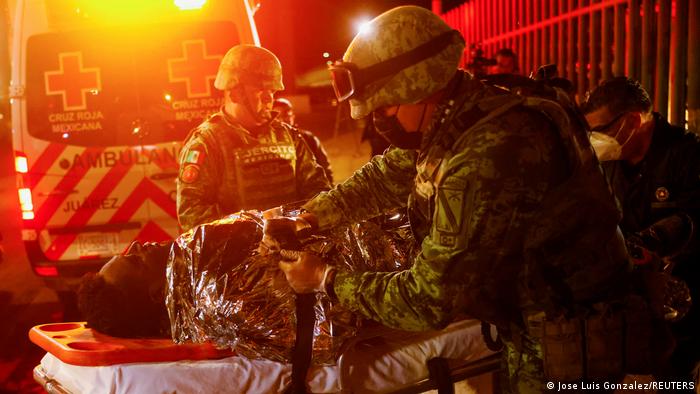 Un militar tapa a una de las víctimas sobre una camilla con una ambulancia y una valla al fondo.