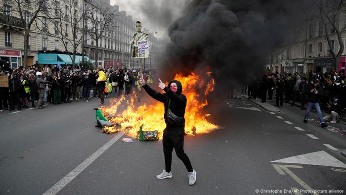 Un manifestante sostiene un cartel burlándose del presidente francés Emmanuel Macron junto a cubos de basura ardiendo durante una manifestación en París. Un manifestante sostiene un cartel burlándose del presidente francés Emmanuel Macron junto a cubos de basura ardiendo durante una manifestación en París.