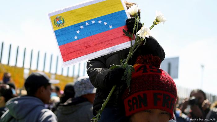 Bei einer Demo hält ein kaum zu sehendes Kind auf den Schultern einer Person Blume und ein Bild der venezolanischen Flagge