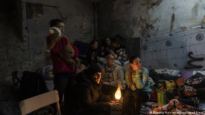 Ein Mann und mehrere Frauen und Kinder sitzen eng beieinander in einem Keller