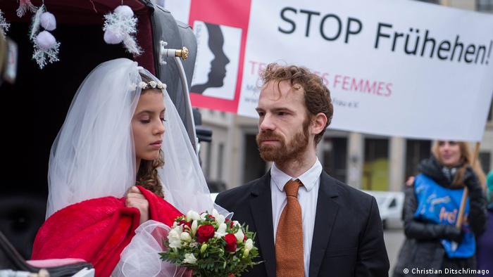 Das Foto zeigt eine Protestaktion der Organisation Terre des femmes 2015 in Berlin. Ein Paar von zwei Erwachsenen, ein Mann und eine Frau, inszenieren sich als Minderjährige, die heiraten. Im Hintergrund fordert ein Plakat STOP Frühehen!.