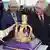 King Charles III looks at a cake of a crown on a purple cushion with other people standing around him