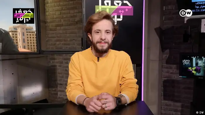 Ali, who is from Syria and transgender, is shown. Ali is in the JaafarTalk studio and is standing behind a black podium. He is wearing a beard and a sweater in a warm shade of orange.