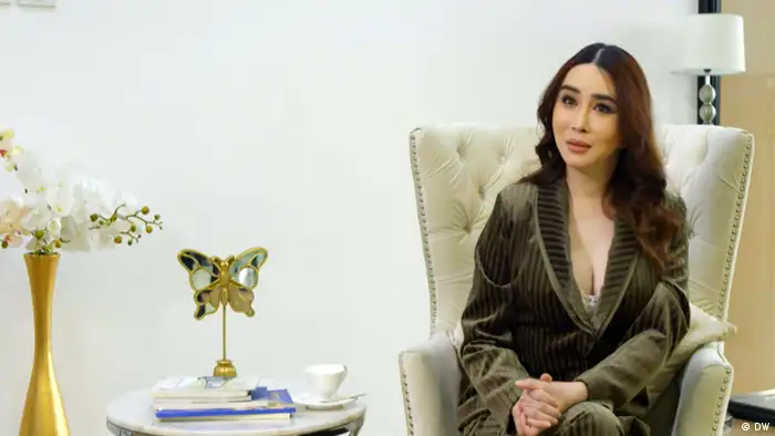 Trans*woman, entrepreneur and mother Anne Jakrajutatip sits on a cream-colored armchair in a hotel room. She wears a dark green velvet suit, and has her hands clasped together. Anne has long brown hair which is slightly wavy.