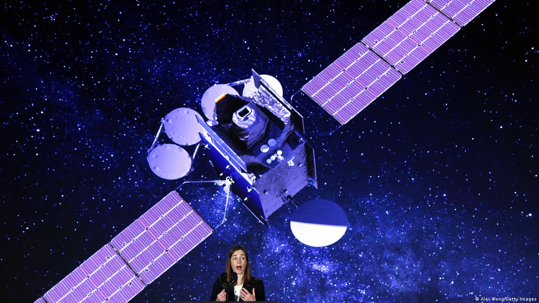 Caroline Nowlan fala em um microfone em frente a um painel com a foto de um satélite