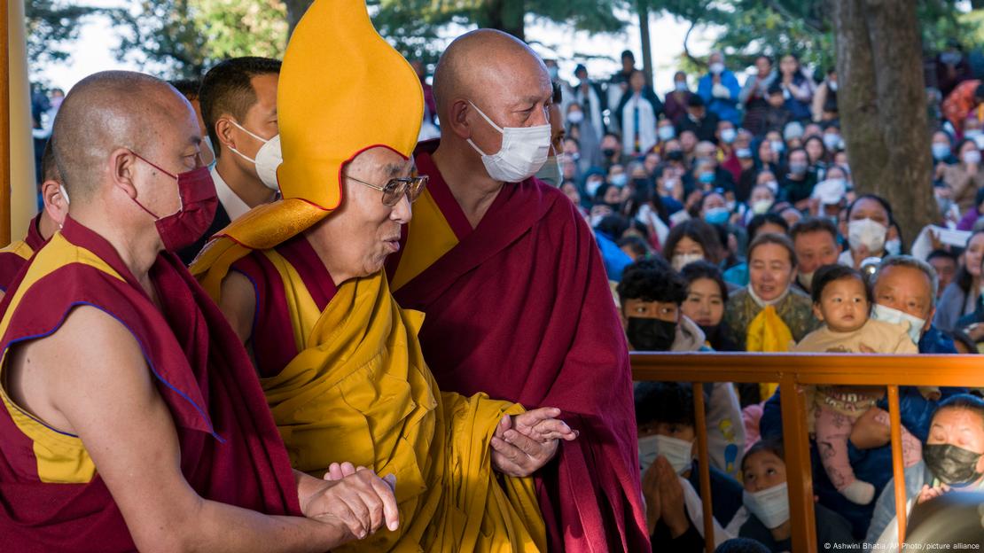 Dalai-lama apoiado em dois ajudantes, com multidão ao fundo