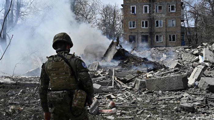 Ein behelmter und mit olivgrüner Schutzweste ausgerüsteter ukrainischer Polizist steht in einem Trümmerfeld zerstörter Häuser