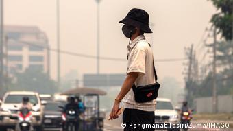 Ein Mann trägt ein weißes Hemd, eine schwarze Maske und einen schwarzen Hut. Er steht auf der Straße. Im Hintergrund sind Autos zu erkennen. 