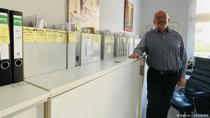 Dirk Jänichen, owner of the Berlin company Jänichen Supply Technology, is standing in front of a shelving cabinet on which there are a number of gray file slipcases and folders. 