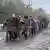 Se ve a un grupo de militares caminando en desordenada formación por una carretera embarrada, el primero de ellos cojeando entre dos compañeros que lo sostienen.