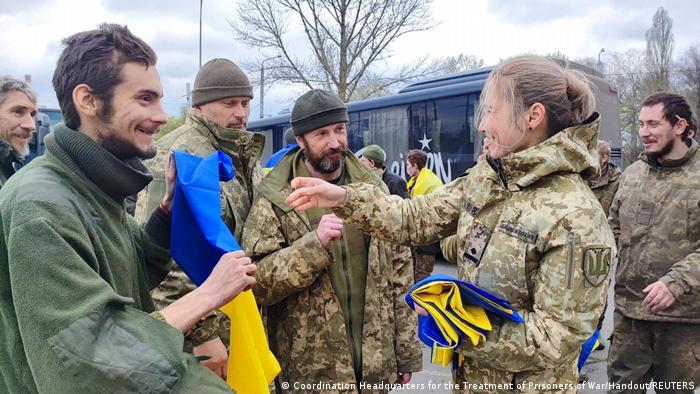 Una soldado entrega banderas ucranianas a los prisioneros liberados entre sonrisas, con un autobús al fondo.