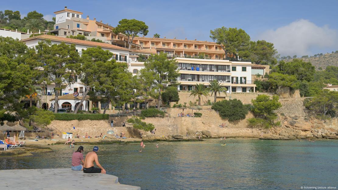 Apartamente pushimi në plazh Platja de Cala Fornells. Shtepi mbi koder buze detit dhe turiste ulur ne breg.
