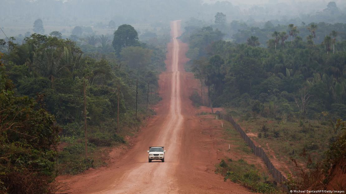 Um carro passa por uma rua de terra que corta a floresta amazônica - é a Transamazônica, na altura de Manicoré, estado do Amazonas. Ao fundo, é possível ver a fumaça de um incêndio. 
