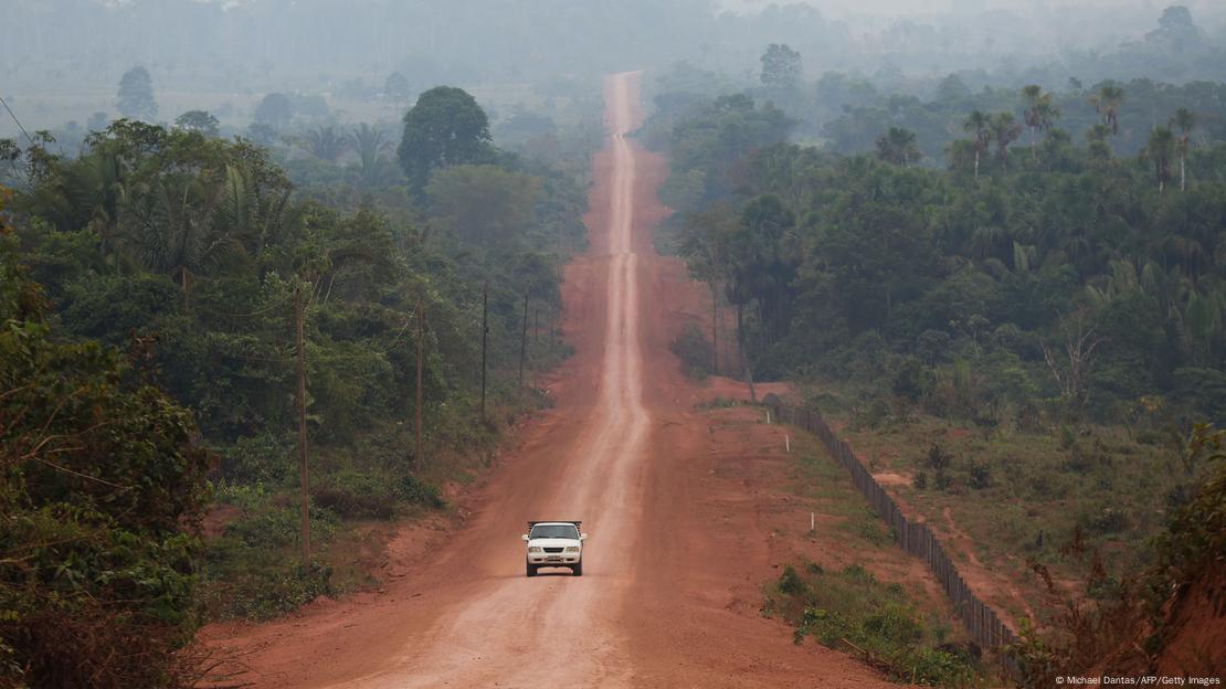 Um carro passa por uma rua de terra que corta a floresta amazônica - é a Transamazônica, na altura de Manicoré, estado do Amazonas. Ao fundo, é possível ver a fumaça de um incêndio. 