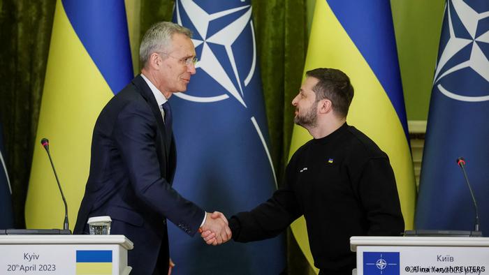 Ein Mann im Anzug und einer im Pullover schütteln sich lächelnd die Hände, im Hintergrund blau-gelbe ukrainische und blaue NATO-Flaggen