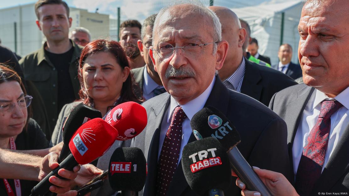 Ο Kemal Kilicdaroglu, άντρας μιλάει σε μικρόφωνα, μια ομάδα ανδρών και γυναικών πίσω του παρακολουθούν και ακούνε.