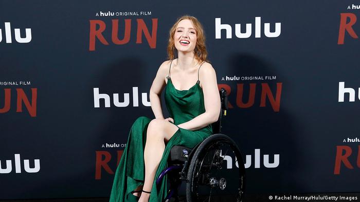 Kiera Allen sitzt in einem grünen Kleid in einem Rollstuhl und lacht.