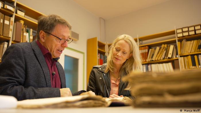 Karin Helmstaedt und Walter Rummel lesen in einem Buch, im Hintergrund sind Bücherregale zu sehen.