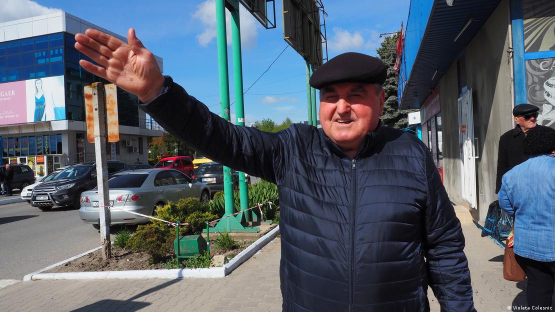 An elderly man stands on the street in the sunshine with one arm outstretched