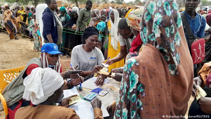Tschad Adre | Sudanesische Flüchtlinge, Welternährungsprogramm: Menschen sitzen an einem Tisch im Freien und bekommen Bescheinigungen, im Hintergrund stehen etliche Personen in weiter Warteschlange