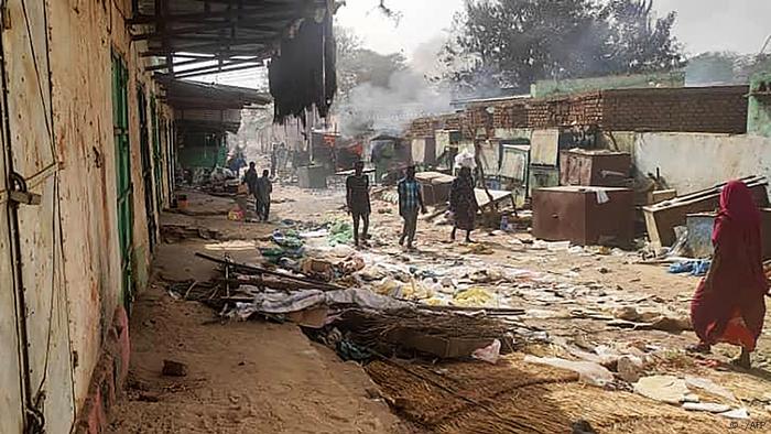 Sudan Konflikt l Kämpfe in Darfur l Markt von El Geneina: Menschen gehen über eine unbefestigte Straße mit Müll und herumliegenden Dingen, im Hintergrund steigt Rauch auf.