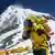 A climber clad in yellow climbing gear and oxygen tanks, against the backdrop of Mount Everest