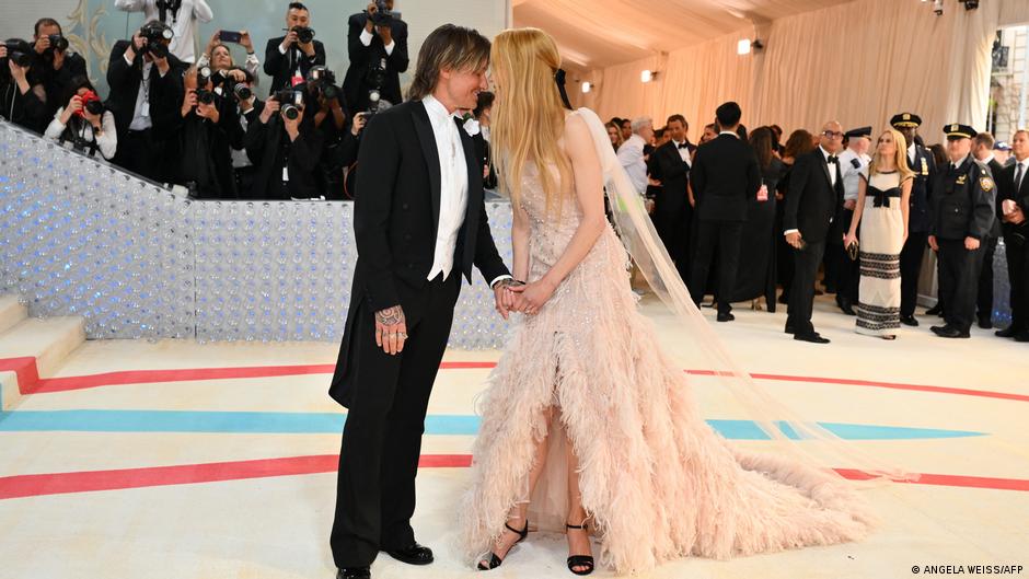 Nicole Kidman in einem weiß-beigen Kleid, das unten mit Federn geschmückt ist, wendet sich ihrem Ehemann Keith Urban zu, der einen schlichten schwarzen Smoking trägt.