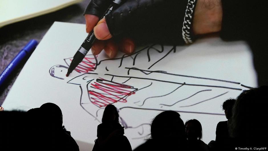 Menschen stehen vor einer Abbildung, die eine Hand des Designers Karl Lagerfeld in einem Lederhandschuh mit offenen Fingerkuppen zeigt. Mit einem Stift fertigt er eine Modeskizze an.