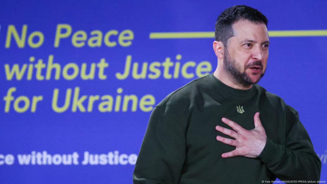 Ukrainian President Volodymyr Zelenskyy holds his left hand to his chest while standing in front of a sign reading 