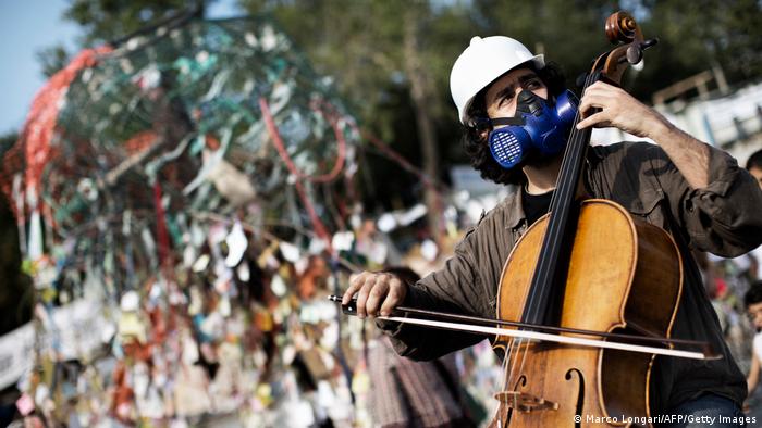 Ein Mann mit Helm und Gasmaske spielt Cello auf einem Platz 