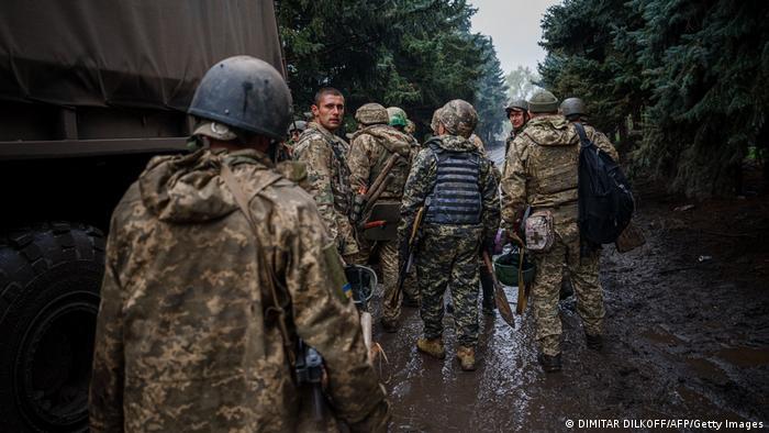 Eine Gruppe ukrainischer Soldaten in Kampfmontur steht mit dem Rücken zur Kamera auf einer Schlamm bedeckten Straße neben einem Militär-LKW