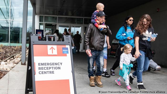 Mehrere Erwachsene mit kleinen Kindern verlassen ein Gebäude. Sie gehen an einem Schild vorbei, auf dem Emergency Reception Centre zu lesen ist.