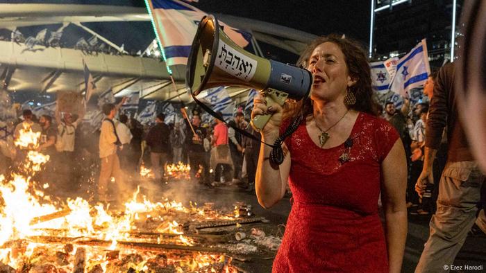 Eine Frau mit Megafon steht auf der Straße, hinter ihr brennt es, im Hintergrund Menschen mit israelischen Fahnen