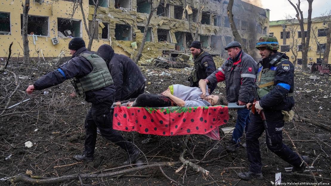 Homens carregam uma mulher grávida em uma maca improvisada, em meio a escombros de prédios