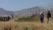Im Hintergrund ist in einer hügeligen Landschaft ein hoher Stacheldrahtzaun zu sehen, der in der Sonne glänzt. Davor gehen und stehen mehrere Personen. Im Hintergrund ist in einer hügeligen Landschaft ein hoher Stacheldrahtzaun zu sehen, der in der Sonne glänzt. Davor gehen und stehen mehrere Personen.