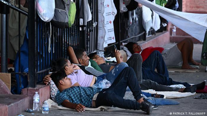 Mehrere junge Männer liegen auf einem Bürgersteig vor einem Zaun, an dem Kleider und Rucksäcke hängen.
