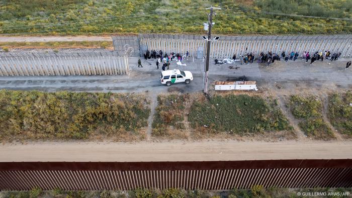 Se ven dos vallas.  Entre ellos hay una pequeña franja de verde.  Junto a ella hay un coche blanco de la guardia fronteriza y una fila de personas.
