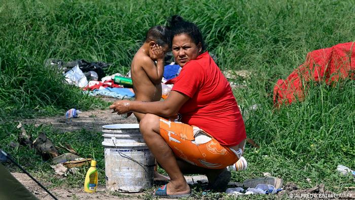 Eine Frau in einem roten T-Shirt blickt in die Kamera, während sie Wasser aus einem Eimer schöpft, um ihr nacktes Kind zu waschen. Die beiden befinden sich auf einer Wiese mit hohem Gras, um sie herum liegen Alltagsgegenstände und Müll. 
